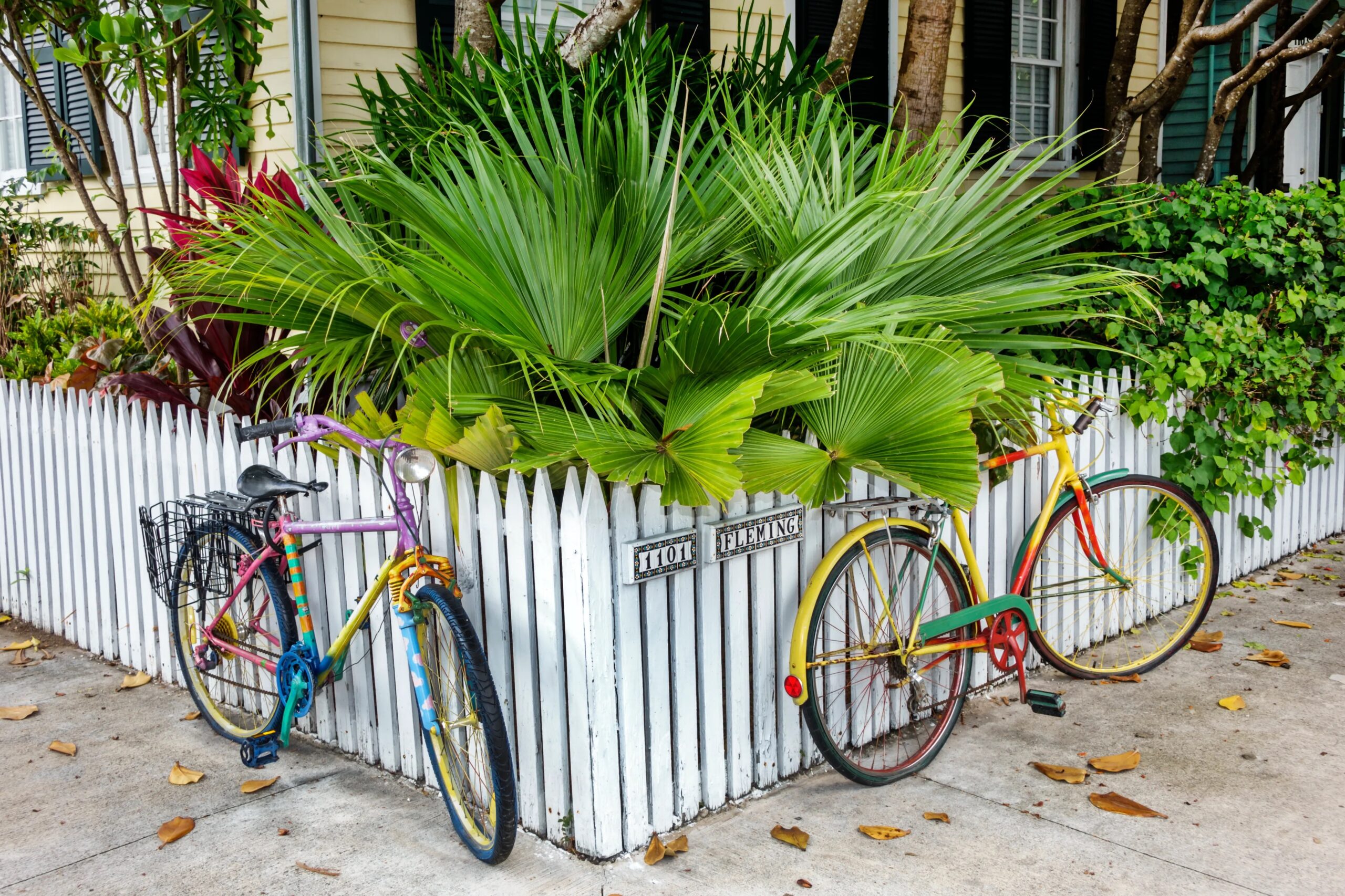 Two bicycles at the corner of a white picket fence with greenery behind the fence.