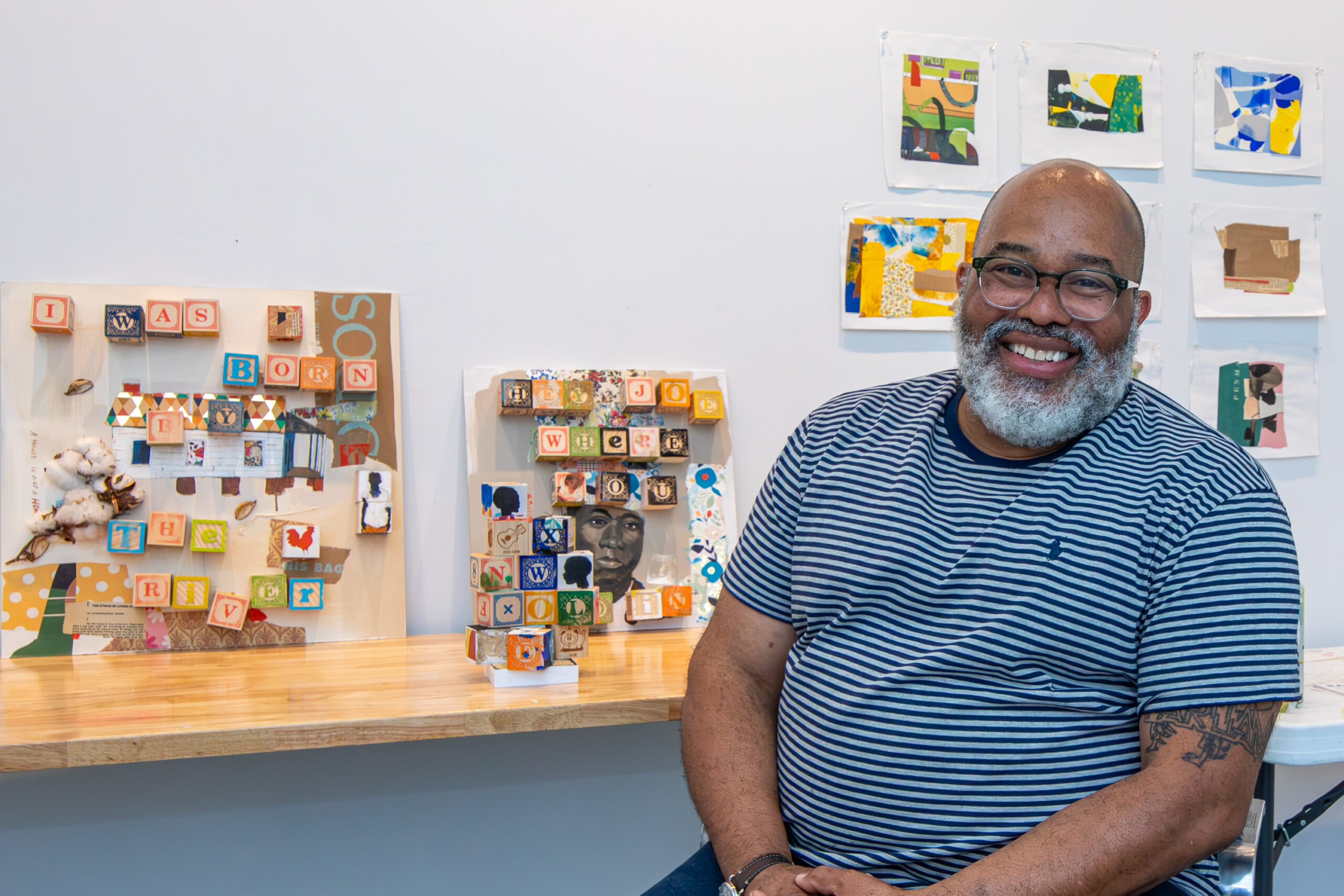 Person sitting at a worktable, smiling with art pinned to the wall behind.