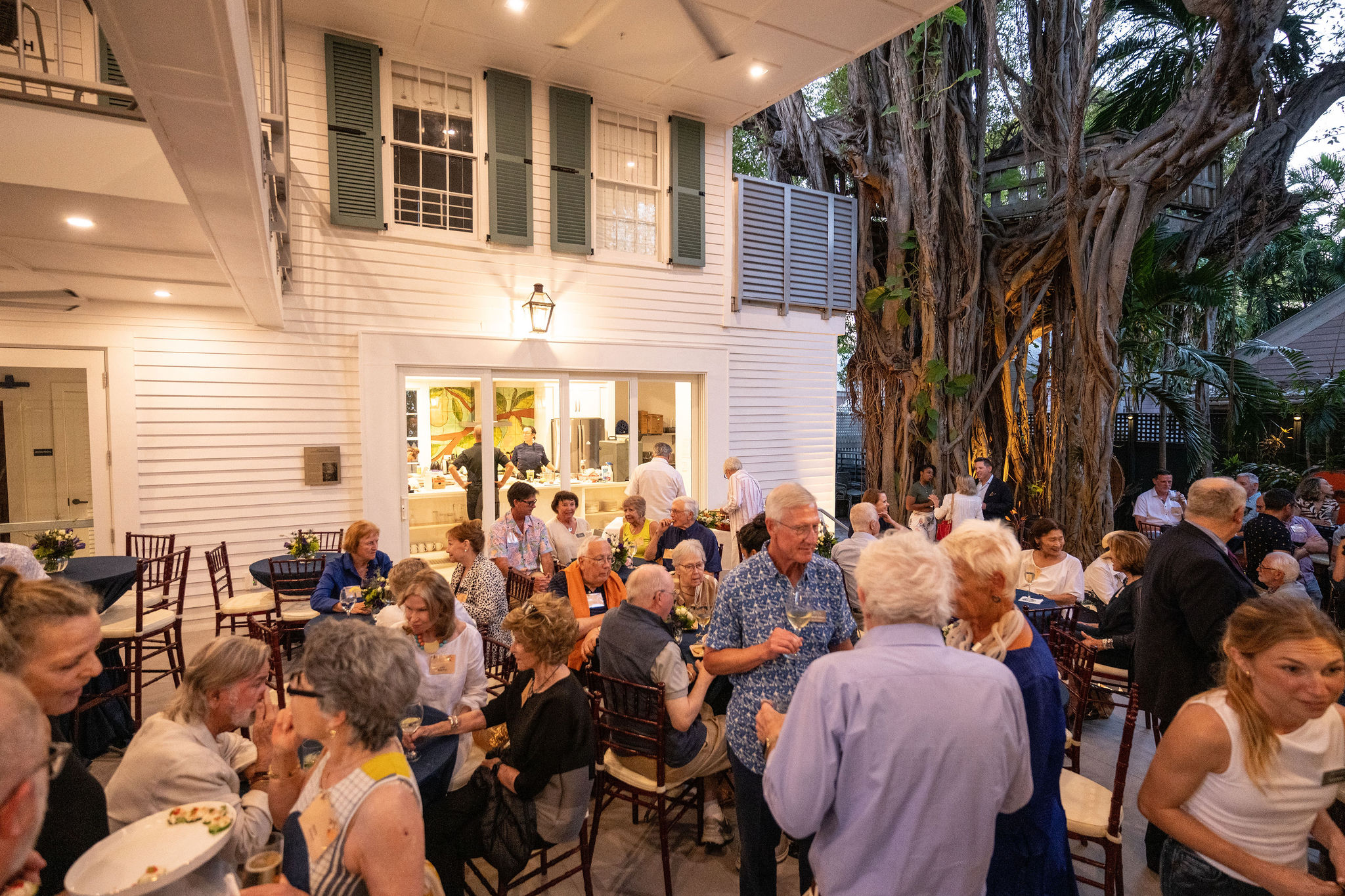 View of a courtyard in the evening with a crowd of people enjoying an event under the covered porch with trees around it.