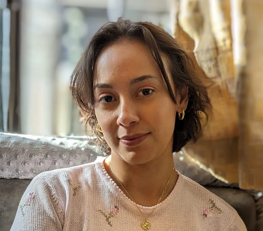 Portrait of a woman with short, wavy brown hair, wearing gold hoop earrings and a delicate gold necklace with a pendant, dressed in a light pink knit sweater with a small floral pattern, looking directly at the camera with a slight smile.