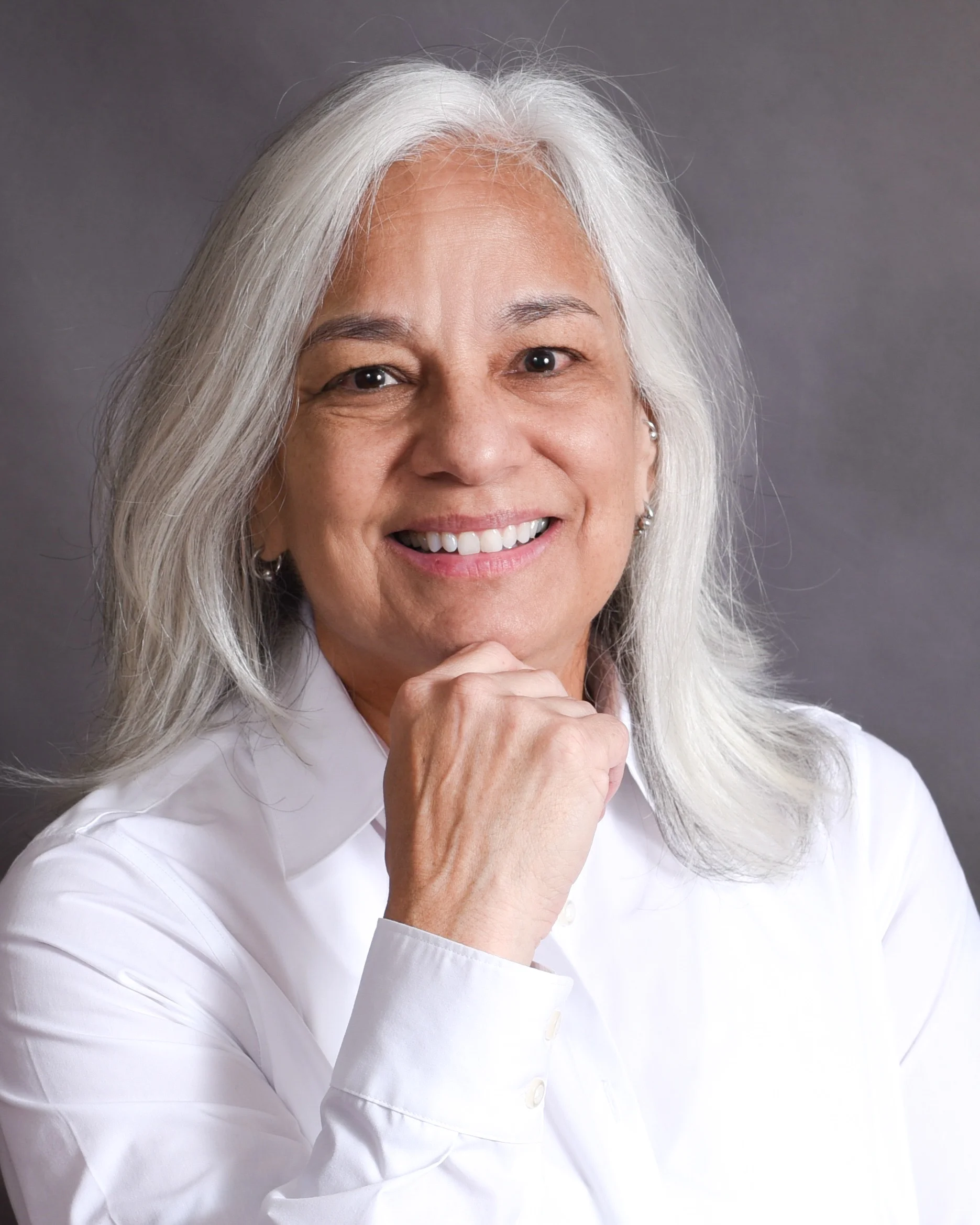 LaDonna BlueEye, performer of Girl of Fire, poses for the camera in front of a grey textured background. She has on a white shirt and has her hand closed and raised as she settles her chin on it.