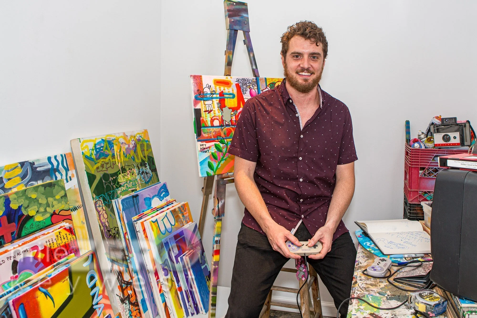 Jack McDonald, a man wearing a maroon polka-dot shirt, sits on a wooden stool holding a video game controller, smiling at the camera in a colorful art studio surrounded by painted canvases, an easel with a mixed-media artwork, and vintage cameras."