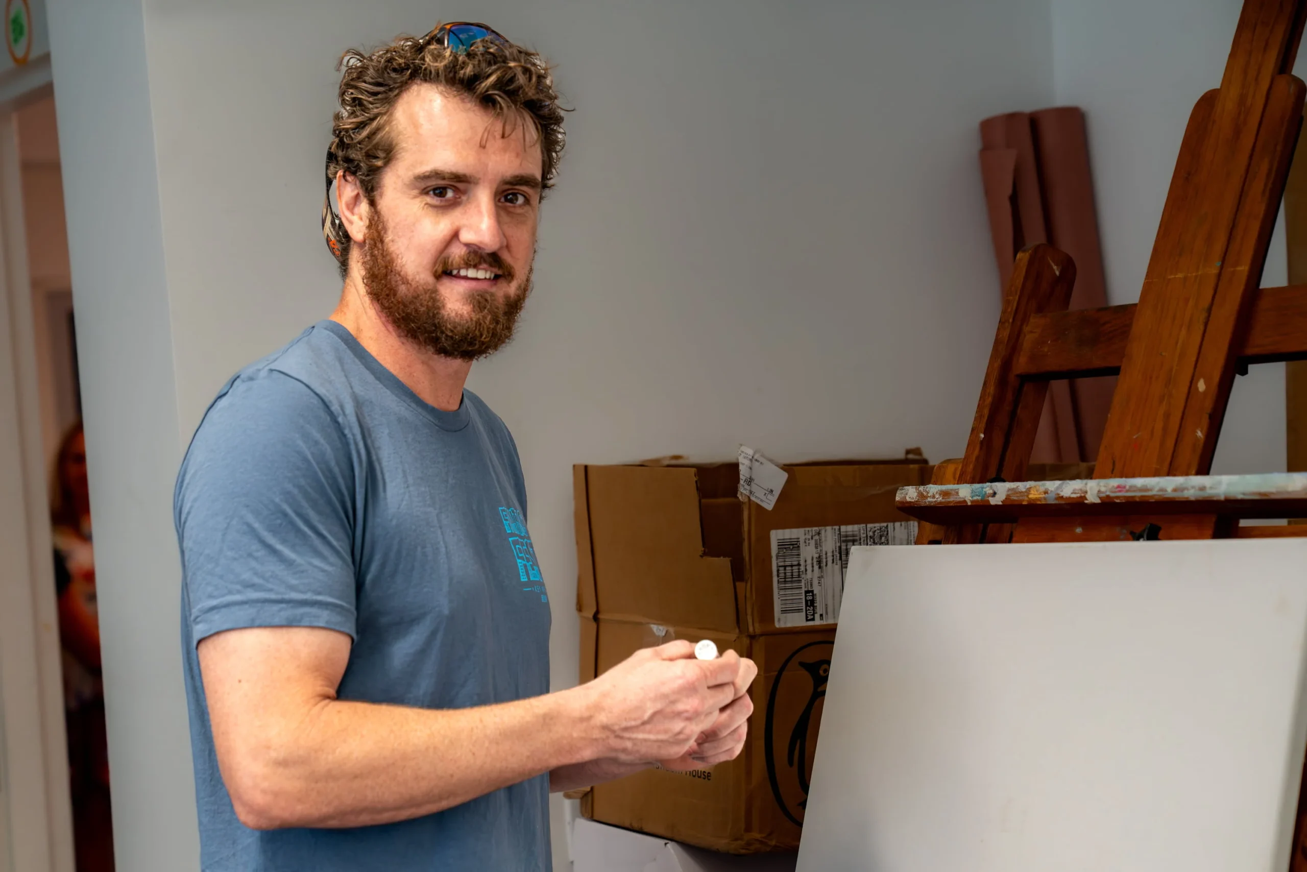 Jack McDonald, a man wearing sunglasses pushed up on his head and a blue t-shirt, stands beside a wooden easel holding a blank canvas, smiling at the camera in a room with cardboard boxes and rolled paper in the background.