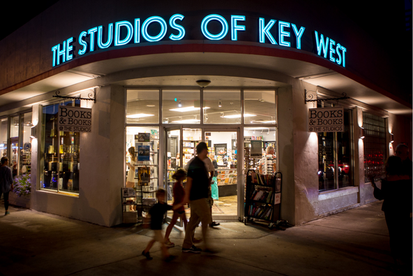 Nighttime exterior of Books & Books at The Studios of Key West, with blue neon lettering above a curved corner entrance and a lit bookstore visible through the glass as people walk past.