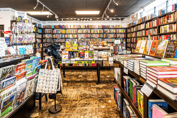 Interior of a bookstore with floor-to-ceiling shelves on all walls, a center table displaying banned books, a magazine rack, tote bags, and printed author names running along the upper wall.