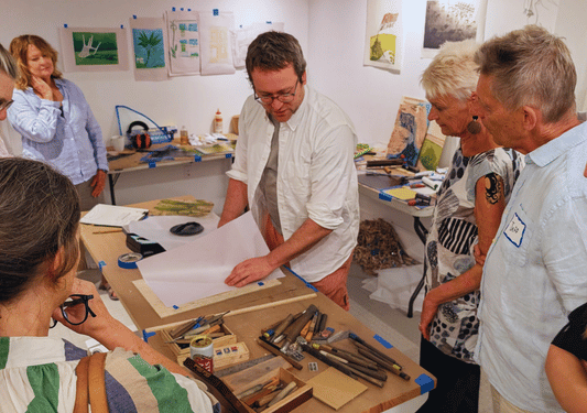 An instructor demonstrates a printmaking technique to a group gathered around a worktable spread with carving tools, prints taped to the wall behind them.