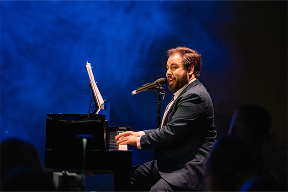A pianist sings into a microphone while playing a grand piano onstage under dramatic blue lighting.