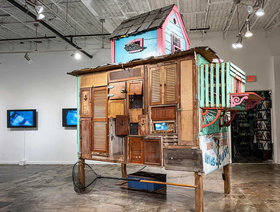 Large sculptural pigeon house installation resembling a colorful, weathered Key West house, constructed from mismatched wooden shutters, doors, and salvaged materials inside a gallery.