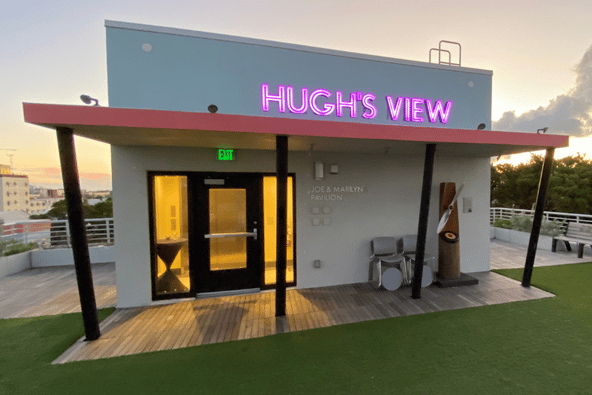 A small rooftop building at dusk with 'Hugh's View' in pink neon above a coral-red awning, glass doors glowing from within, surrounded by wood decking and artificial turf.