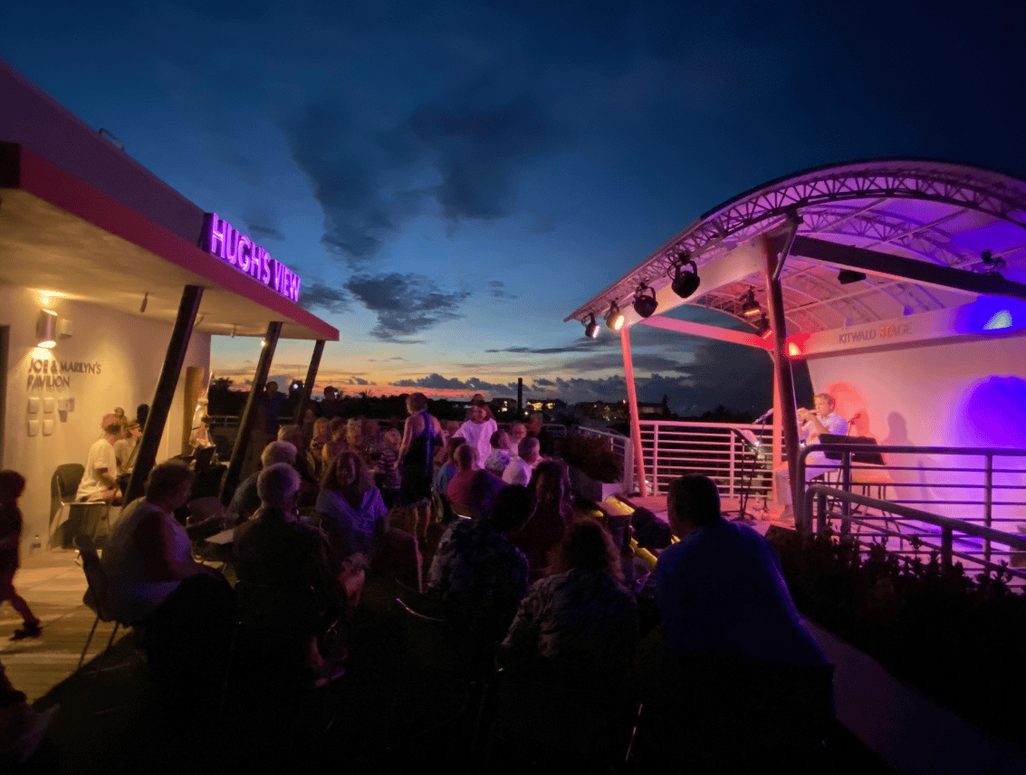 An outdoor rooftop venue at dusk, where a seated crowd watches a performer on a curved canopy stage lit in pink and purple, beneath a deep blue sky with an orange sunset at the horizon.