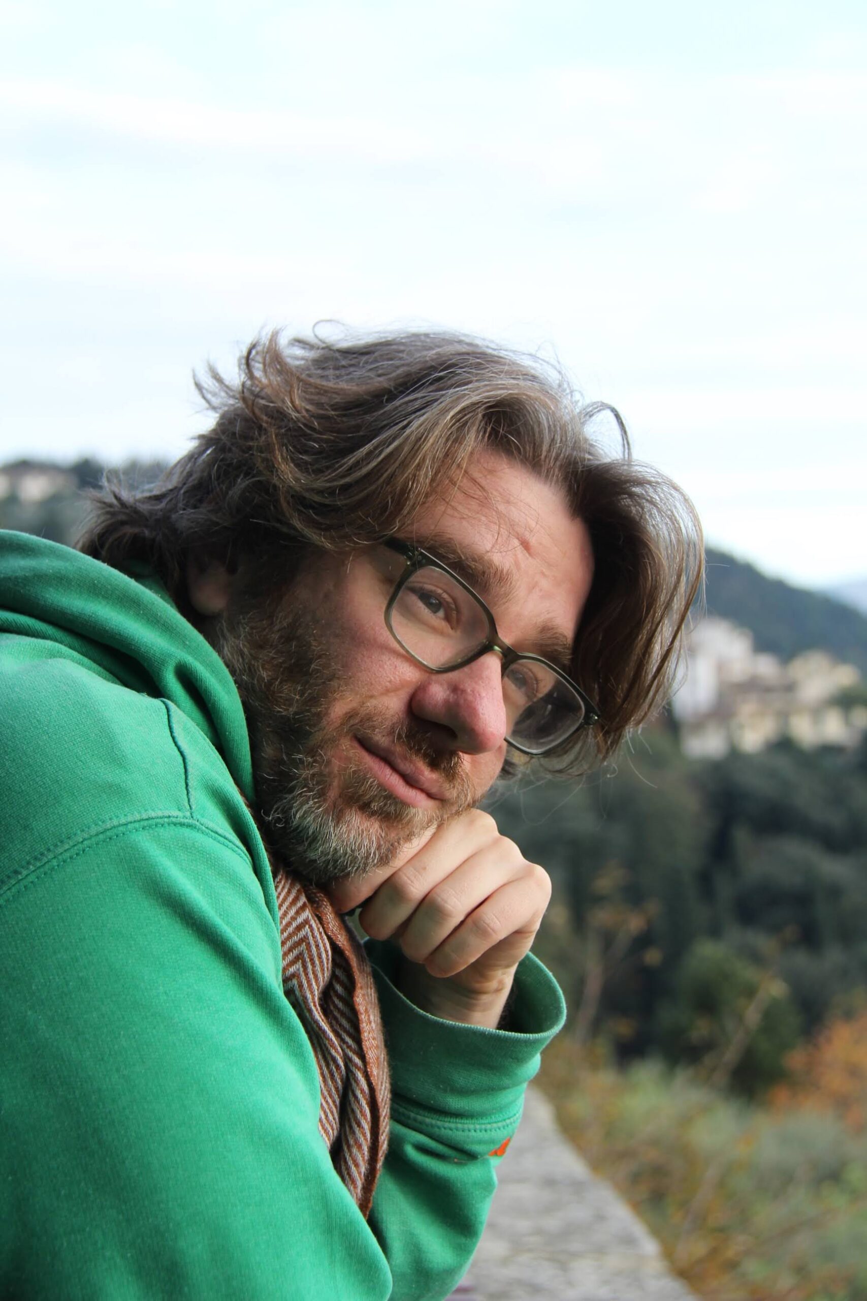 Portrait of Dan McCabe wearing glasses and a green hoodie, leaning on his hand with a wooded hillside in the background.
