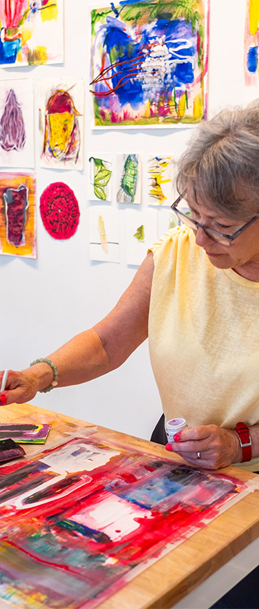 Artist Jan Blythe works at a table surrounded by colorful drawings and paintings during her residency at the PEAR House.
