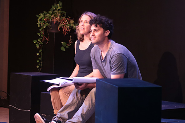 Two actors sit closely together onstage, looking upward during a theatrical performance.