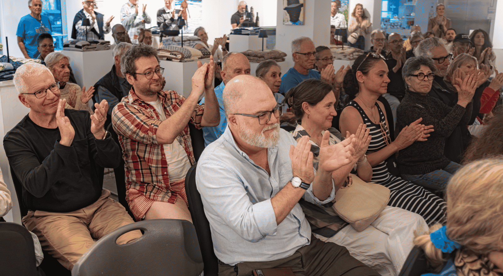 An audience of about two dozen adults seated in folding chairs, applauding at an event held in a bright gallery space displaying folded textiles and framed artwork.