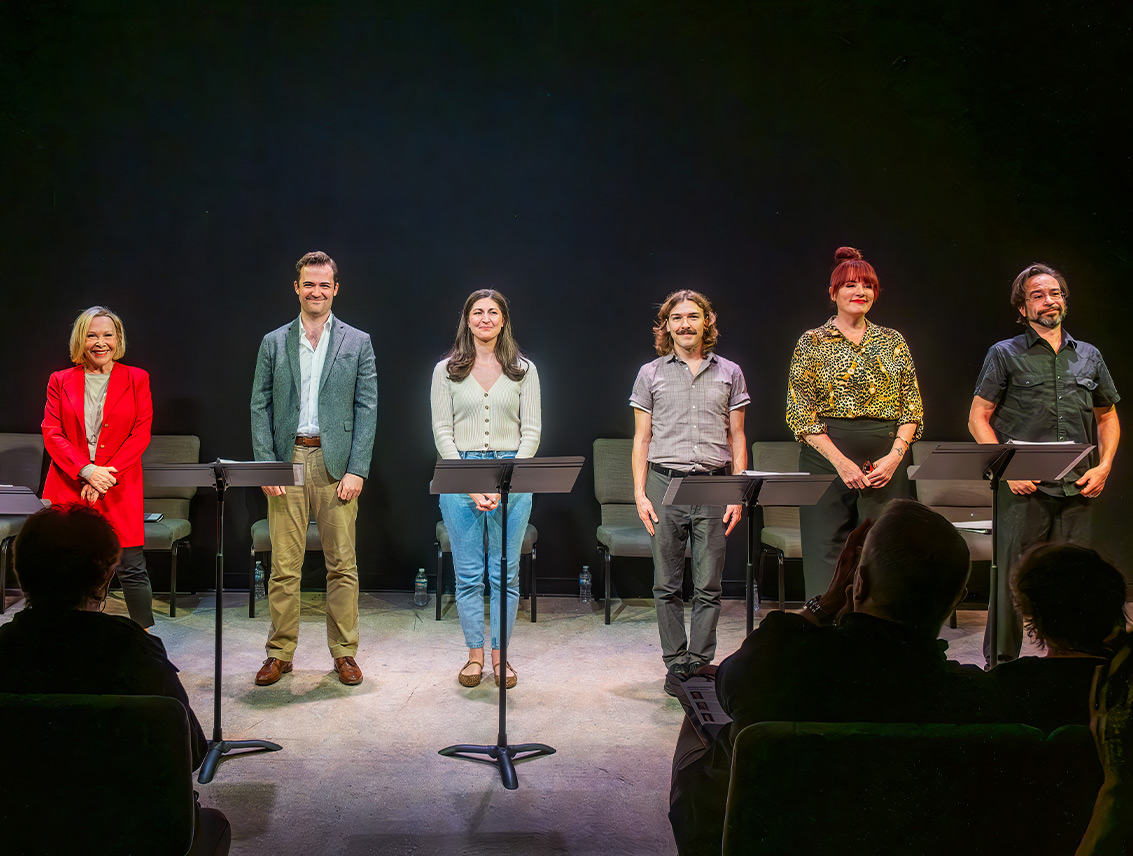 Six actors stand at music stands facing the audience at the end of a staged reading.