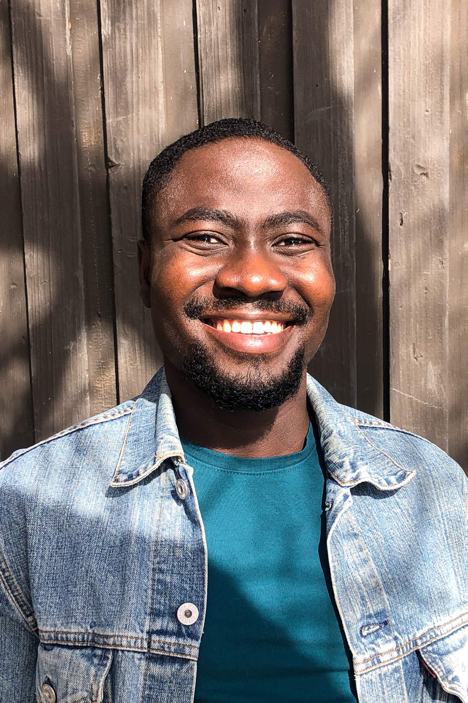 Portrait of a smiling man wearing a denim jacket and teal shirt, standing in bright sunlight against a wooden wall.