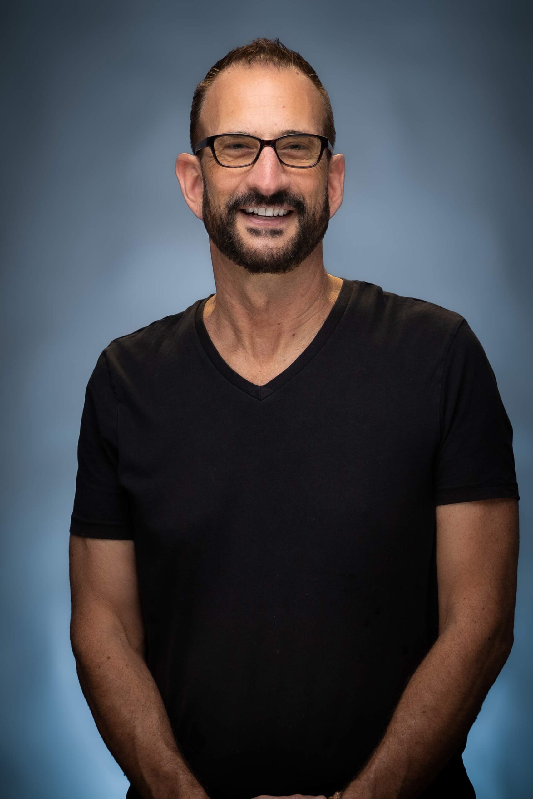 Portrait of Andy smiling in a black T-shirt and glasses against a blue studio background.