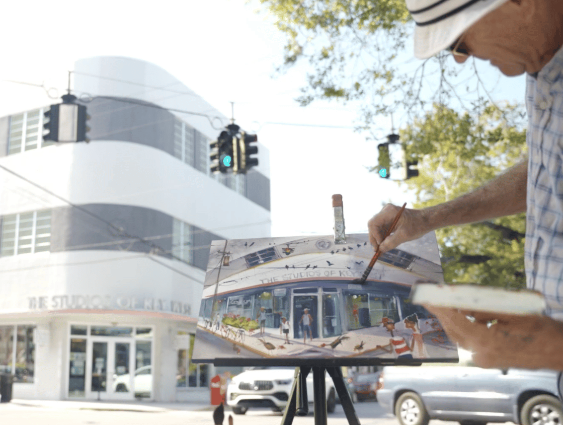 A painter works at an easel on the sidewalk, capturing the curved Art Deco facade of The Studios of Key West that stands behind him.
