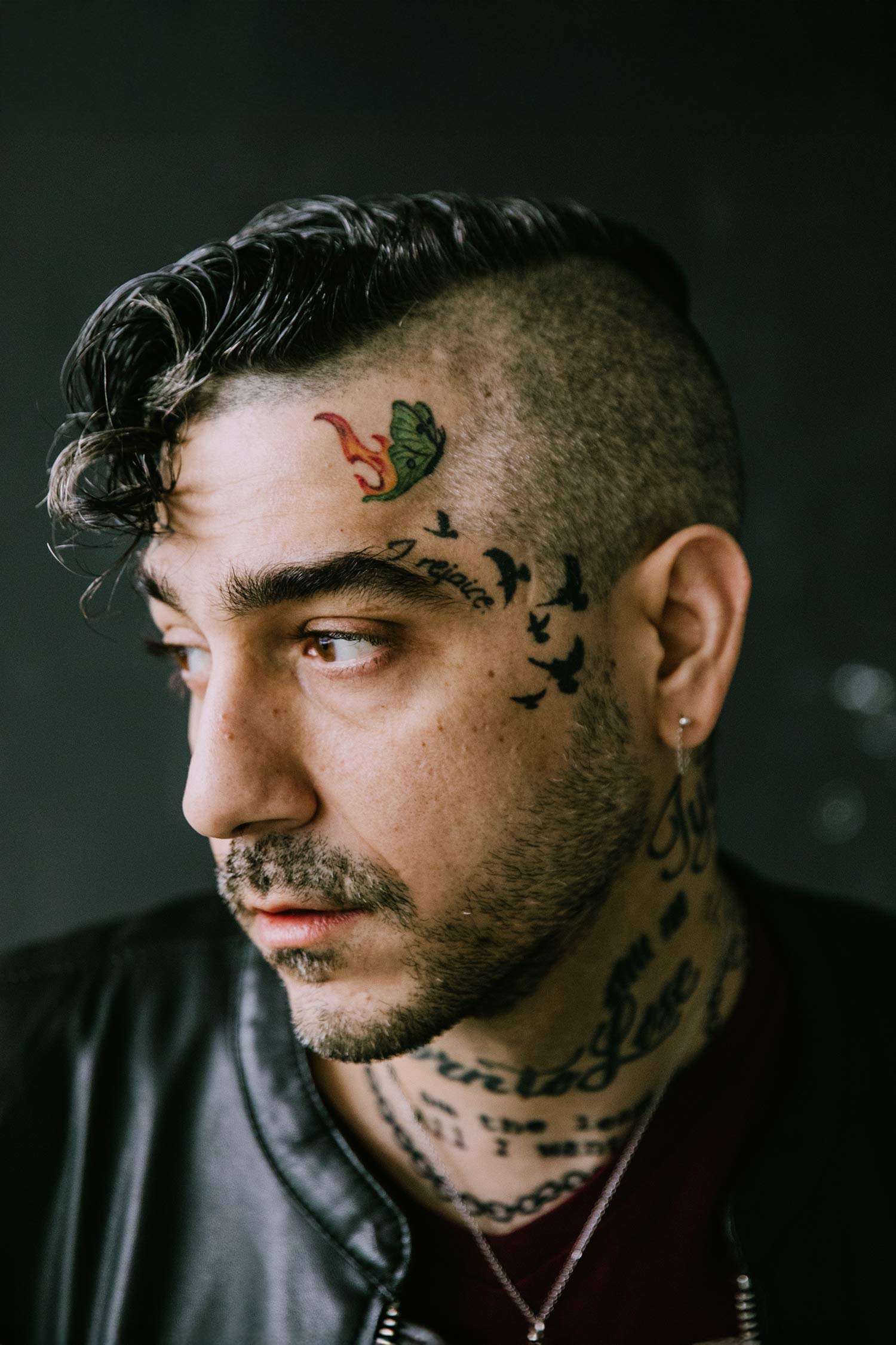 Close-up portrait of a person with dark hair, facial and neck tattoos, and a black jacket, looking off to the side.