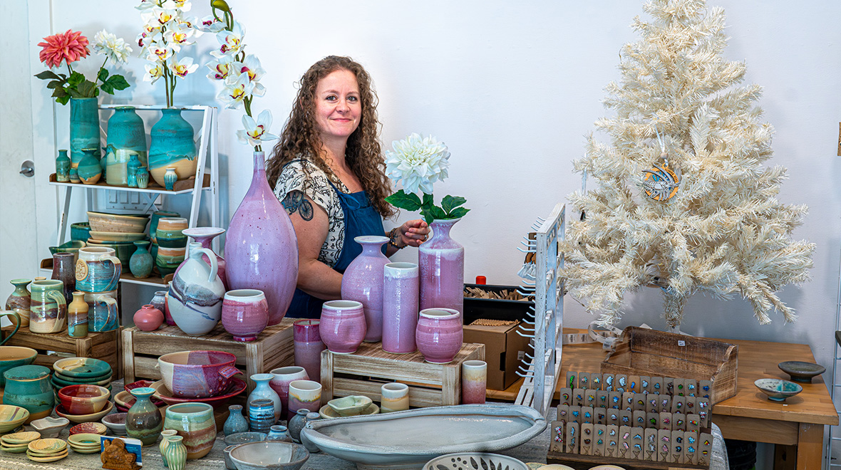 Artist stands behind a colorful display of handmade pottery, ceramics, and jewelry at The Studios’ Art Market.