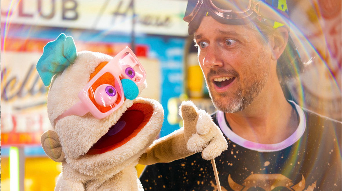 Puppeteer from AstroYeti performs with a playful white puppet wearing oversized pink glasses and a blue nose in a colorful, brightly lit setting.