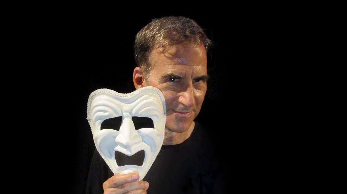 Rich Orloff holds a white theatrical mask beside his face against a black background.