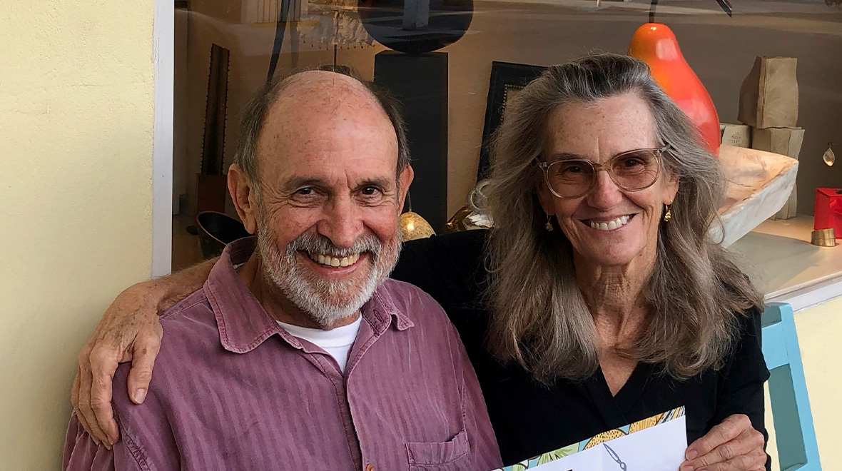 A man and woman smiling together with their arms around each other in front of a gallery window displaying artwork.