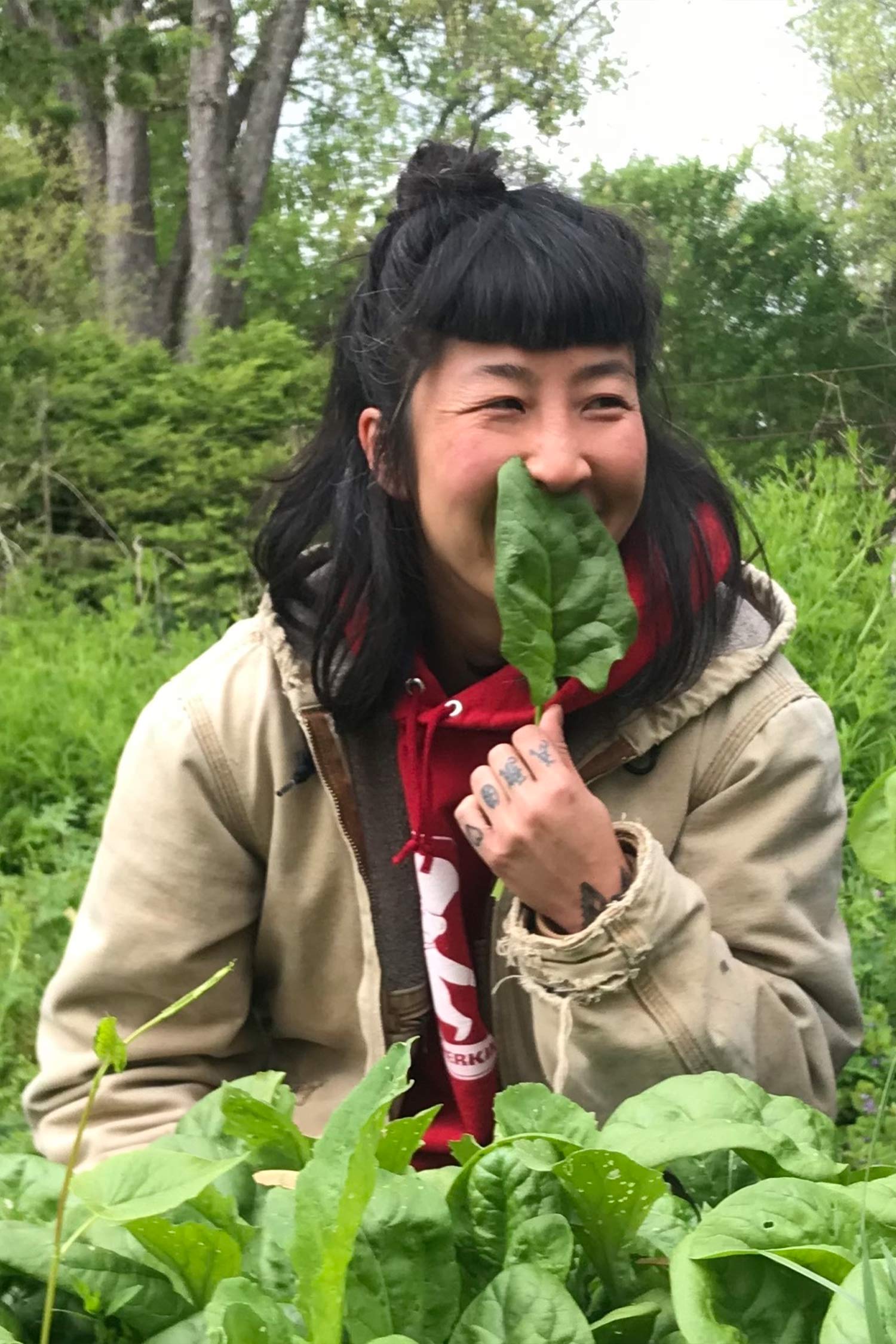 Portrait of a smiling woman seated among lush green plants, holding a large leaf playfully in front of her face.