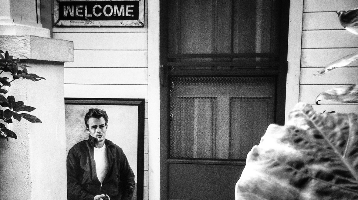 Black-and-white photograph of a framed portrait of James Dean leaning beside the entrance of a house, beneath a “Welcome” sign.