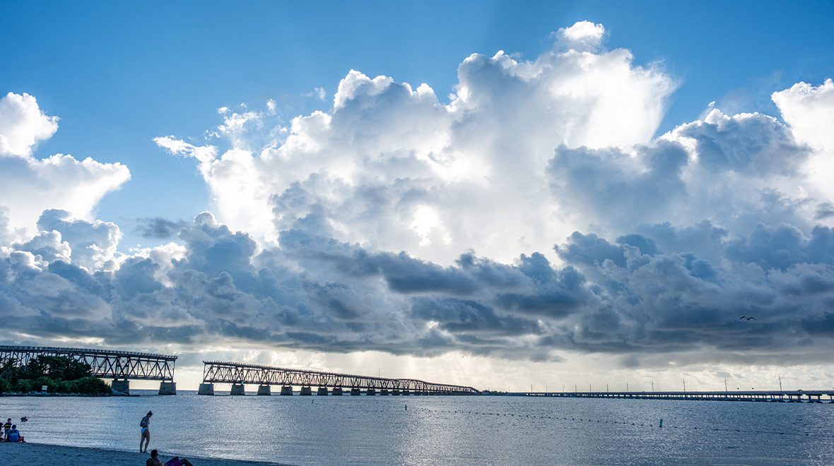 Photograph of the Florida Keys Overseas Railroad bridge beneath towering sunlit clouds, with calm blue water stretching across the foreground.