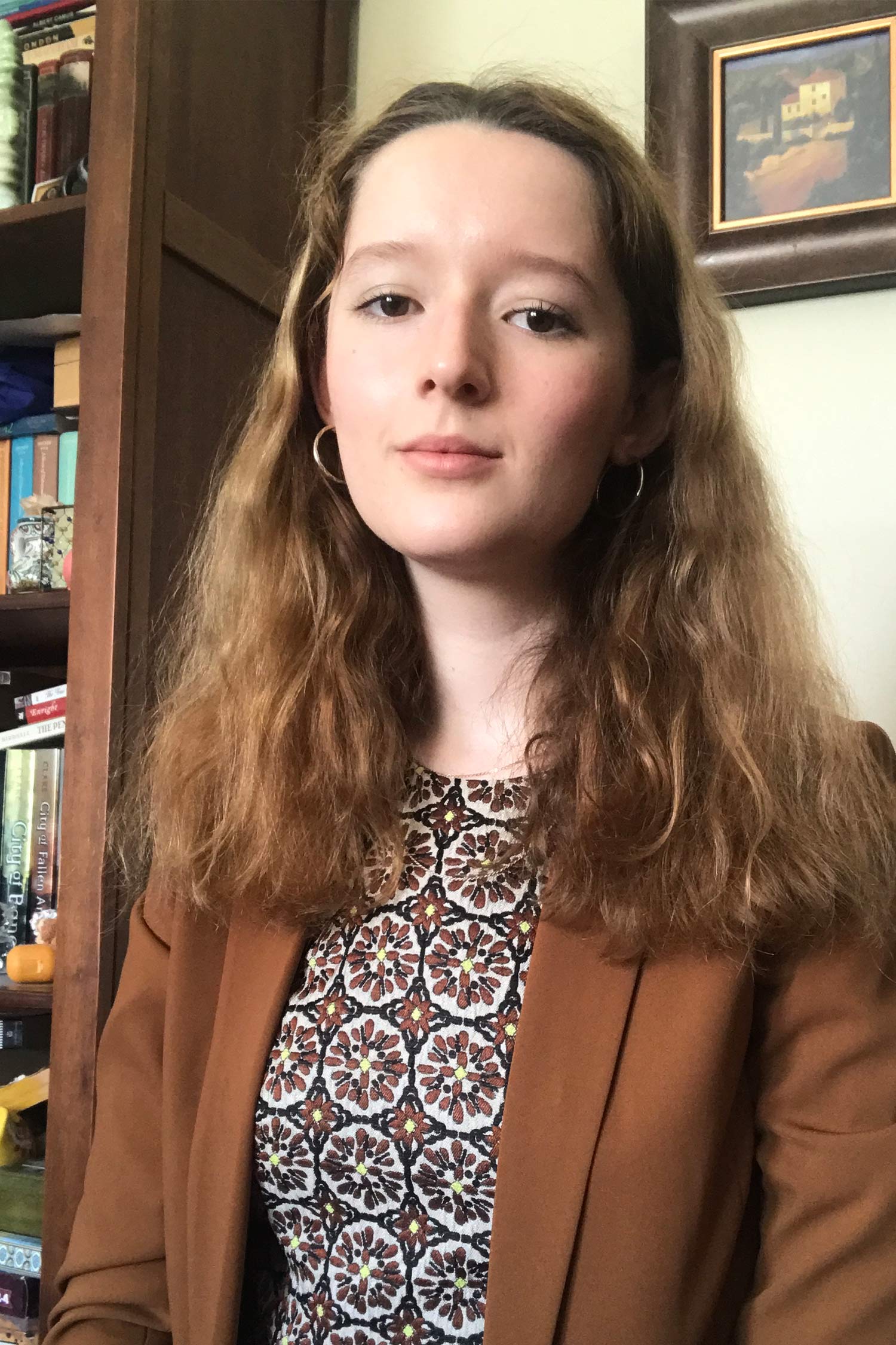 Portrait of a person with long brown hair wearing a brown blazer and patterned top, seated indoors beside a bookshelf.