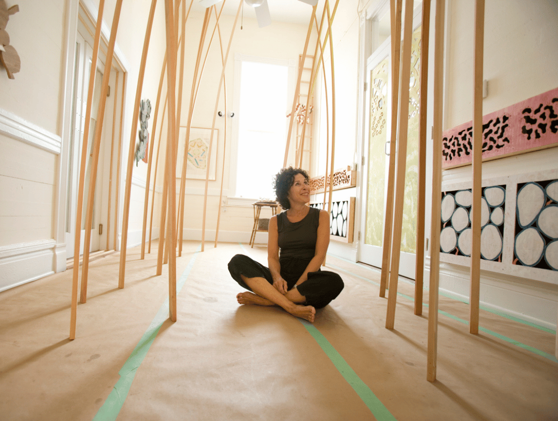 Early studio artist Deb Goldman sits cross-legged on the floor of a sunlit studio, surrounded by tall curving wooden rods that arch overhead, with patterned sculptural panels mounted along the walls.