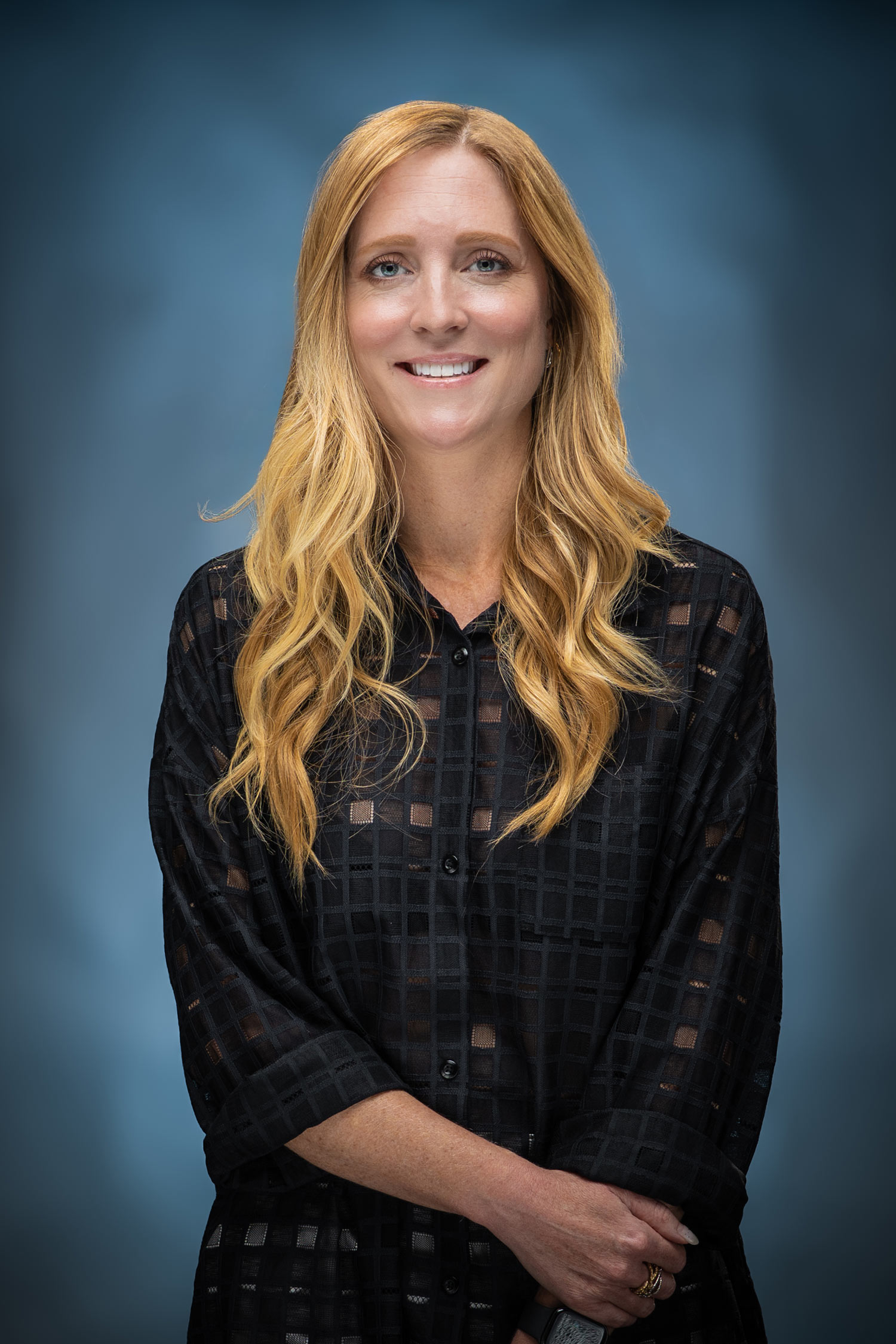 Smiling woman with long strawberry-blonde hair wearing a black patterned blouse, photographed against a blue studio background.
