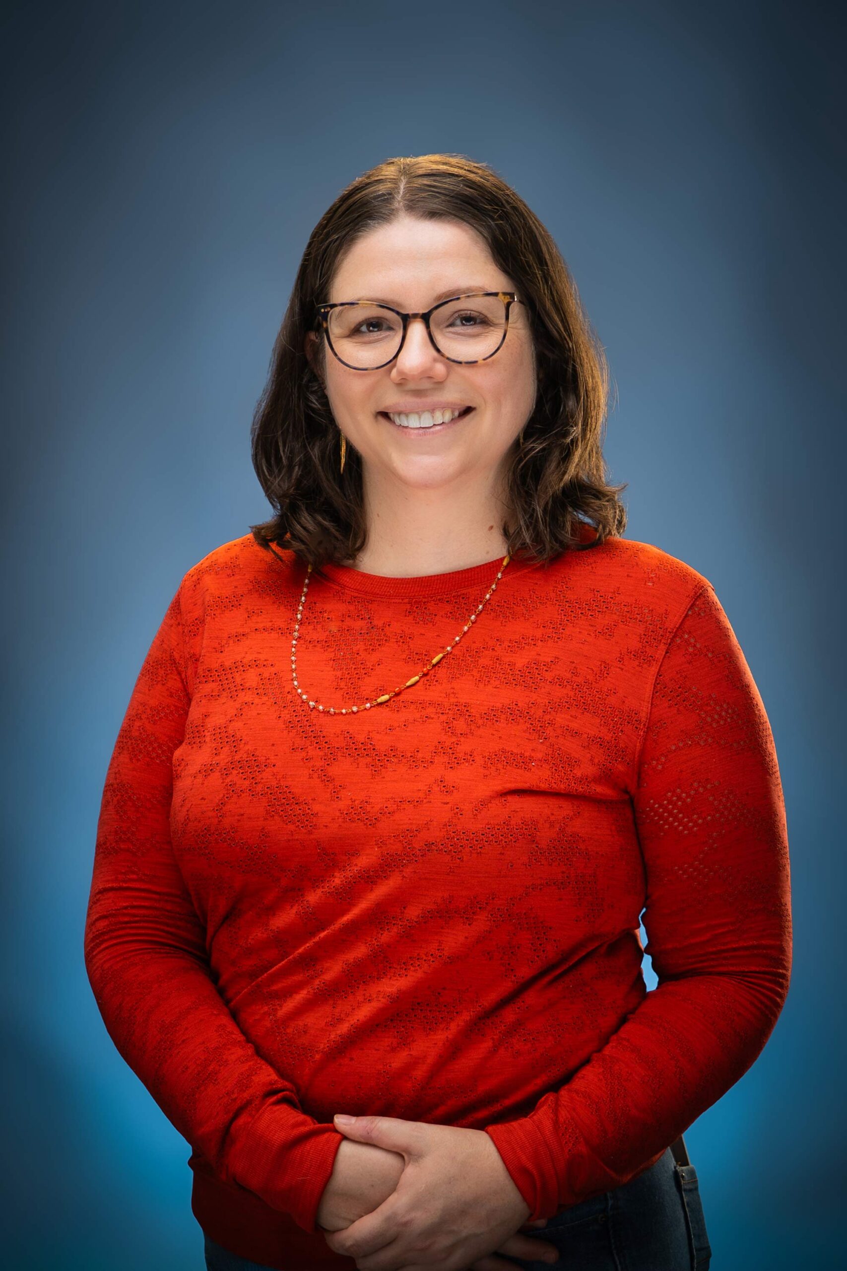 Headshot of Emily smiling in glasses and a red top against a blue studio background.