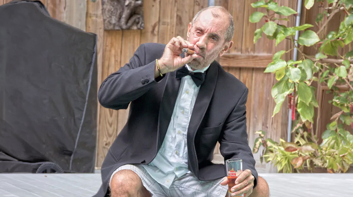 Man in a tuxedo jacket and bow tie sits outdoors, smoking a cigar and holding a drink.
