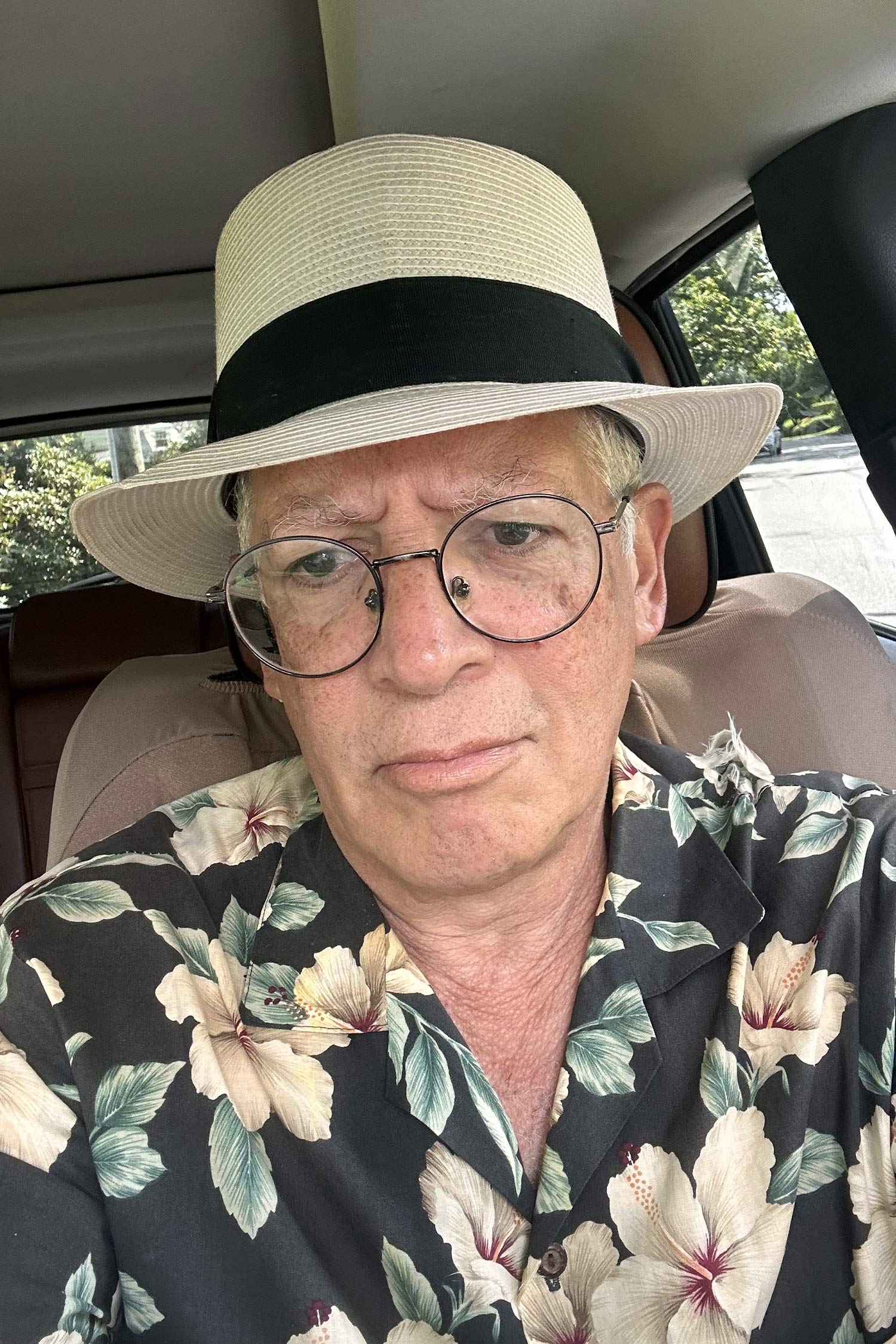 Man wearing round glasses, a wide-brimmed straw hat and a dark floral shirt, seated inside a car.