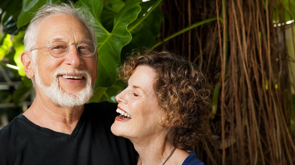 Older couple smiling and laughing together outdoors, surrounded by lush tropical foliage.