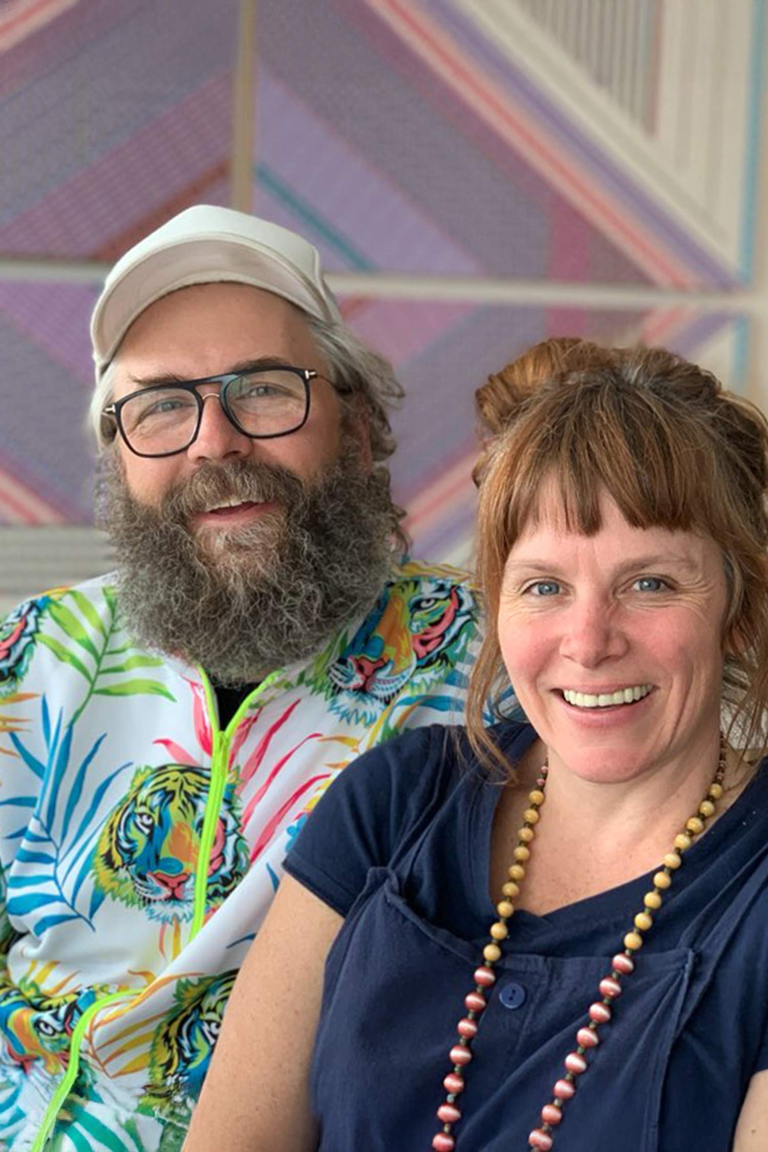 Portrait of two smiling artists seated together, one wearing glasses, a white cap, and a colorful tiger-print jacket, the other wearing a dark top and beaded necklace.
