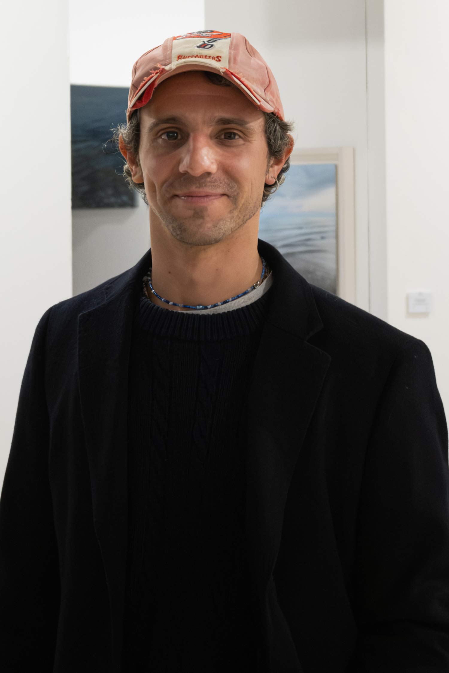 Portrait of a man wearing a faded red baseball cap, black sweater and dark jacket, standing in a gallery with framed artwork visible behind him.