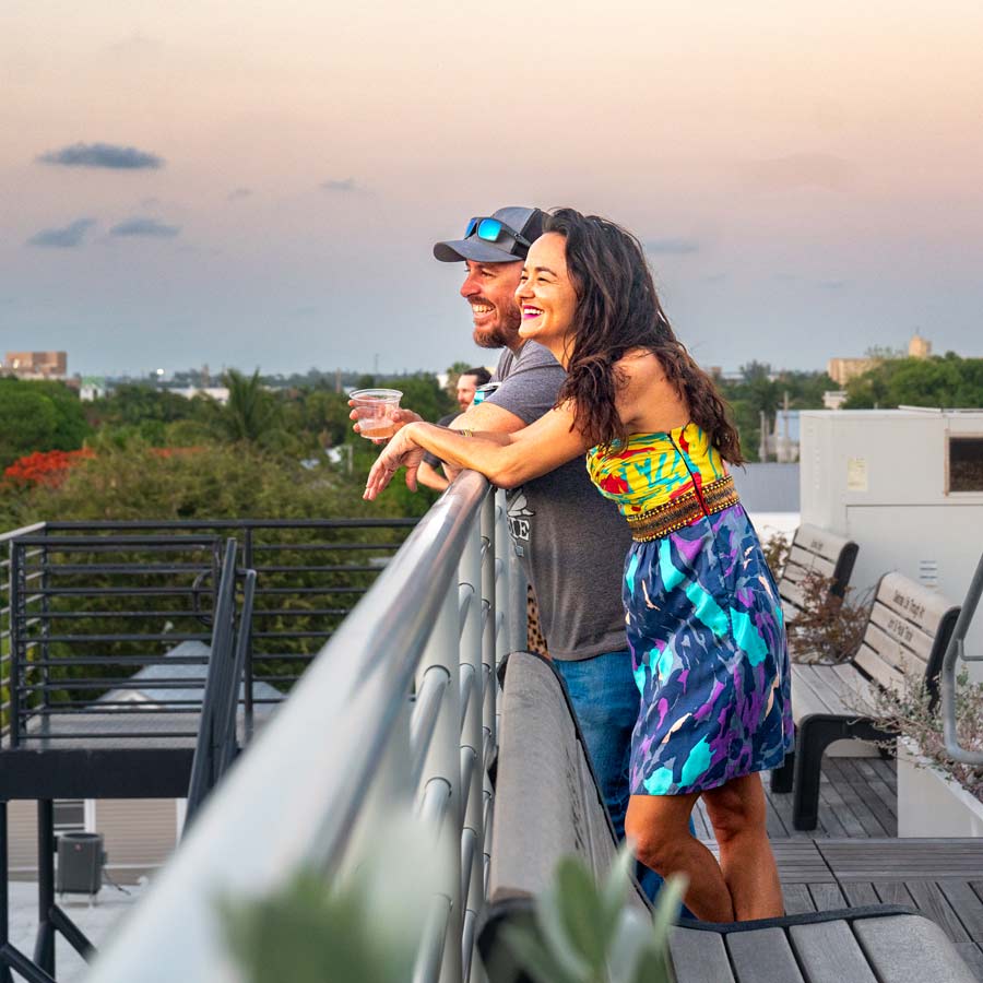 Two guests smile and lean against the railing at Hugh’s View, overlooking the treetops and rooftops of Key West at sunset.