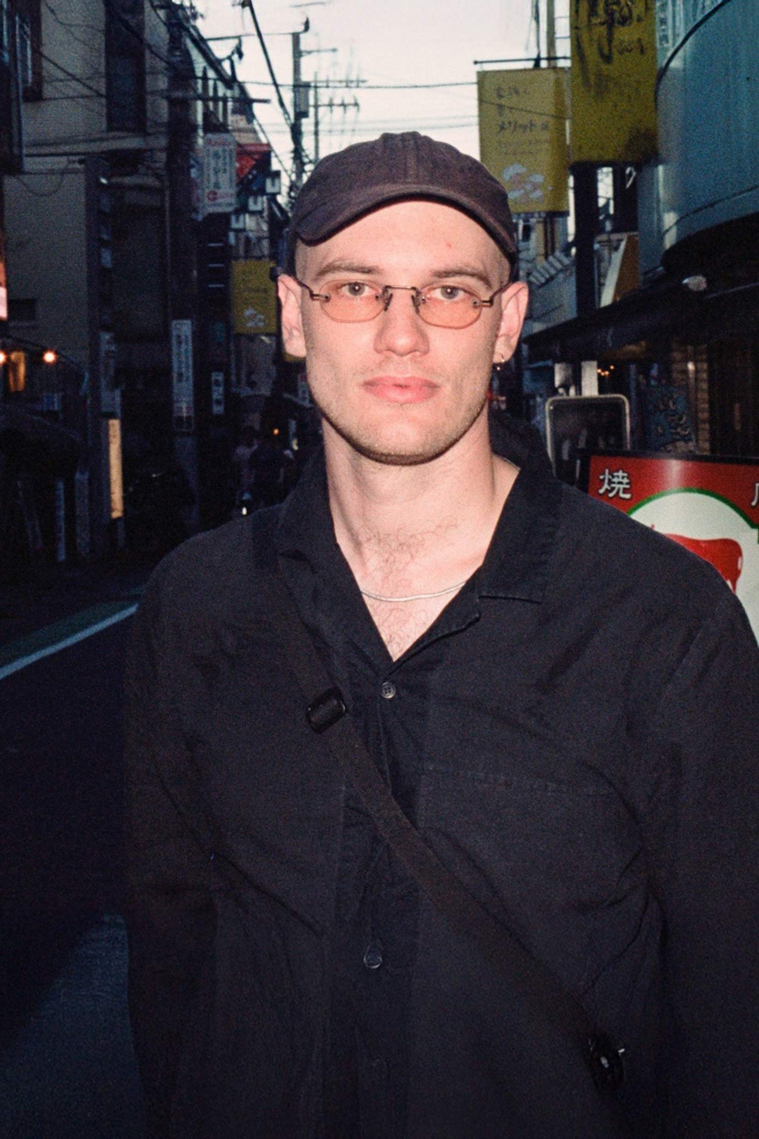 Connor Greer stands on a narrow city street wearing glasses, a dark cap, and black shirt.