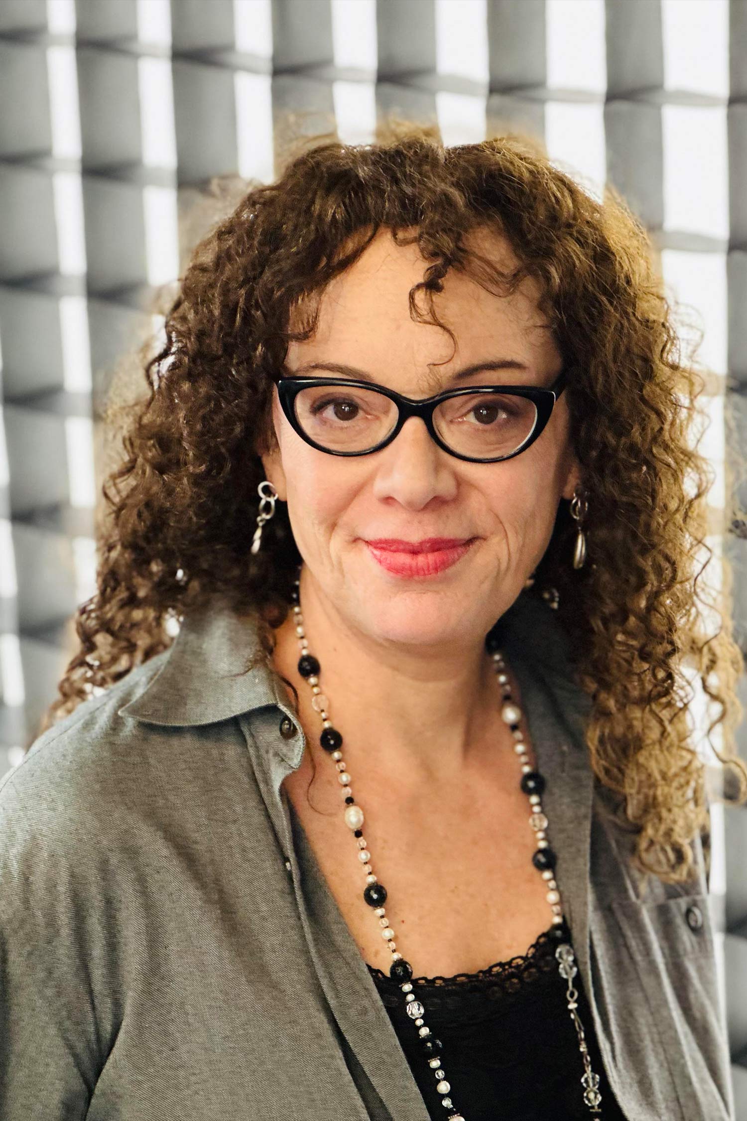 Portrait of a woman with long curly hair and black glasses, wearing a gray shirt and beaded necklace against a softly patterned background.