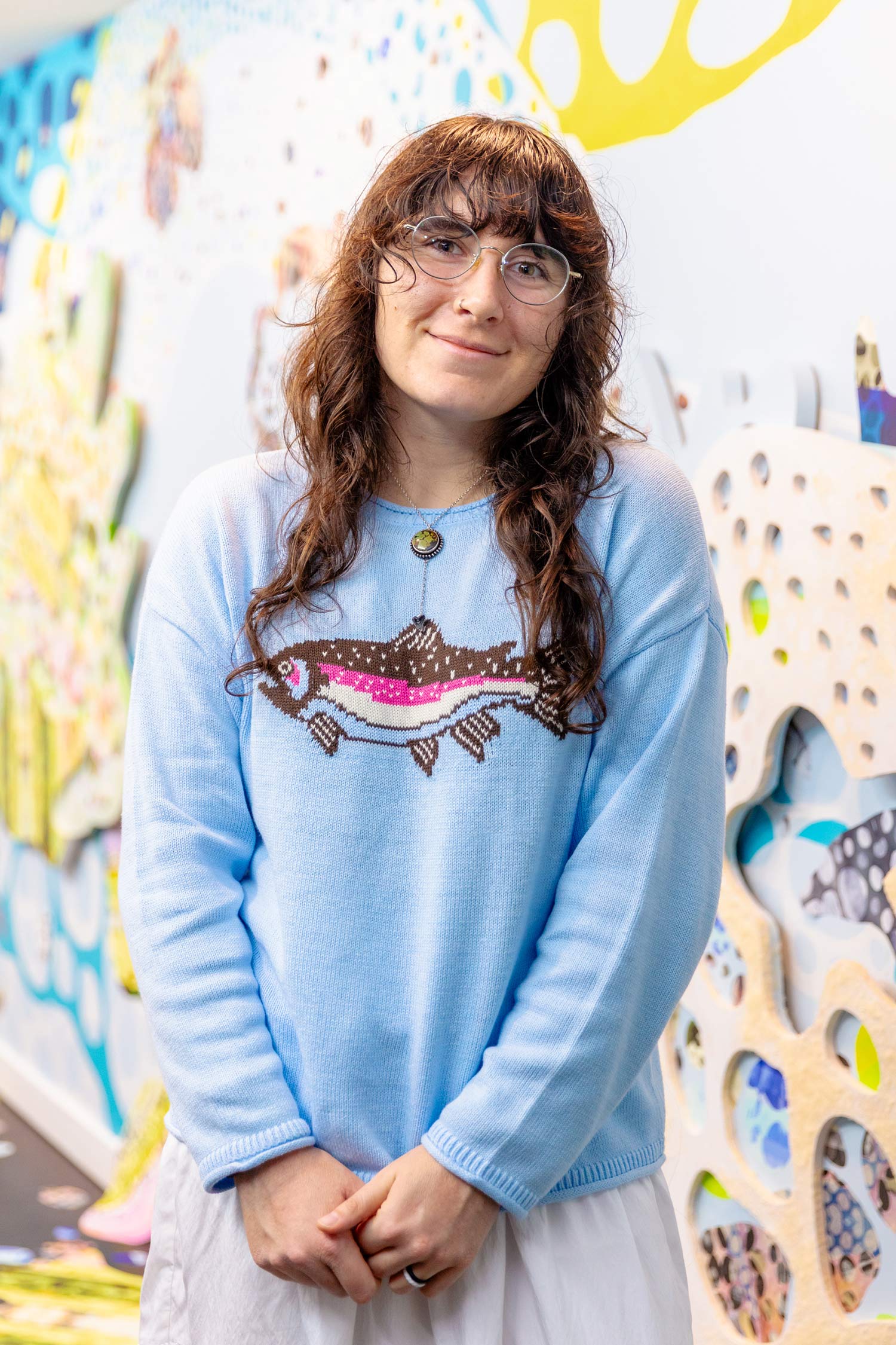Portrait of Hannah Keats smiling in front of her colorful installation, wearing glasses and a light blue sweater with a fish design.