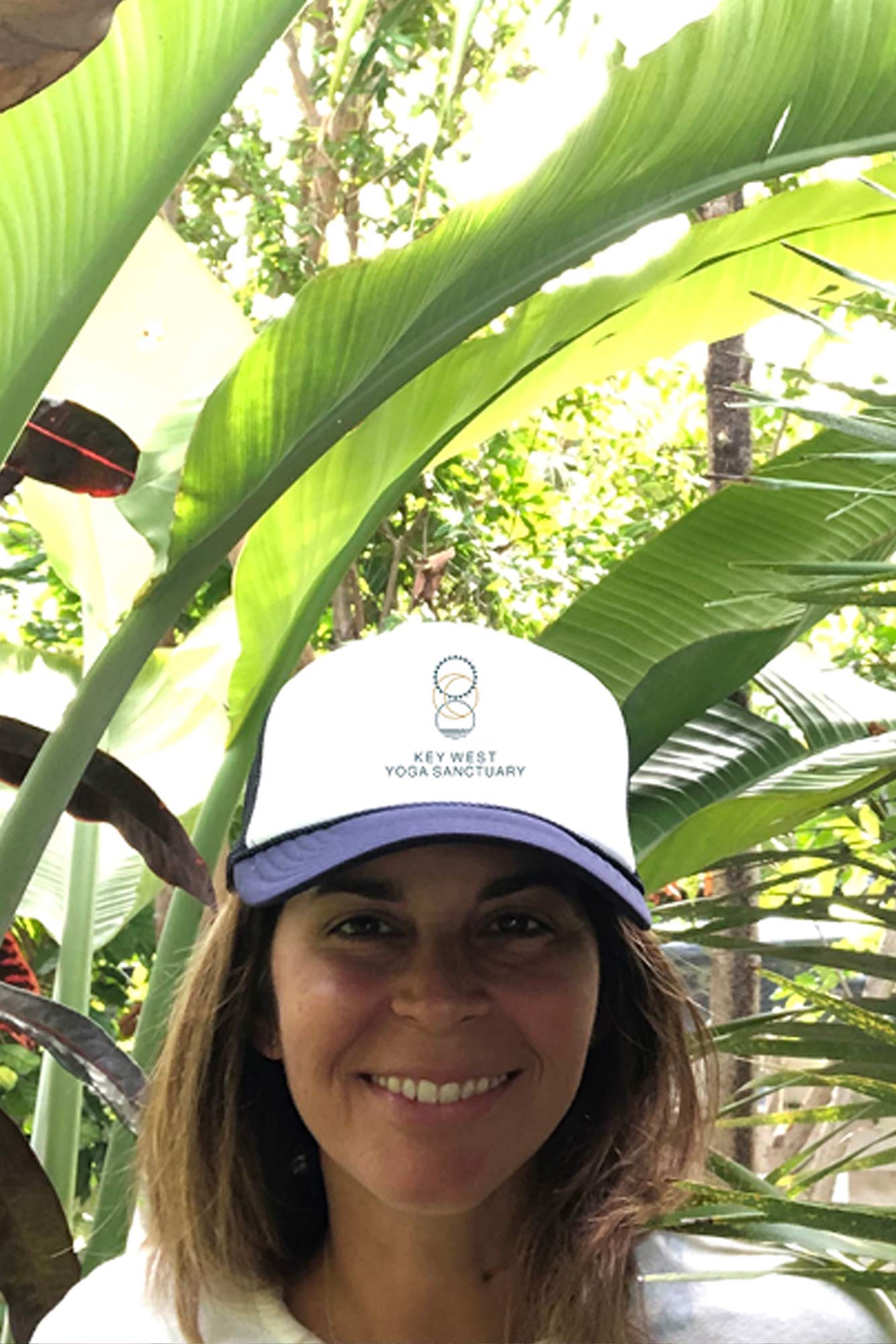 Erika Hawks smiling outdoors in a Key West Yoga Sanctuary hat, surrounded by lush tropical foliage.