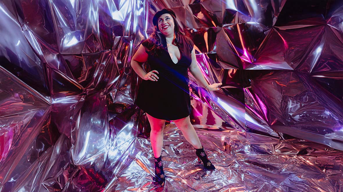 A woman in a black dress and heeled ankle boots poses amid crinkled metallic walls reflecting vivid pink, purple and blue light.