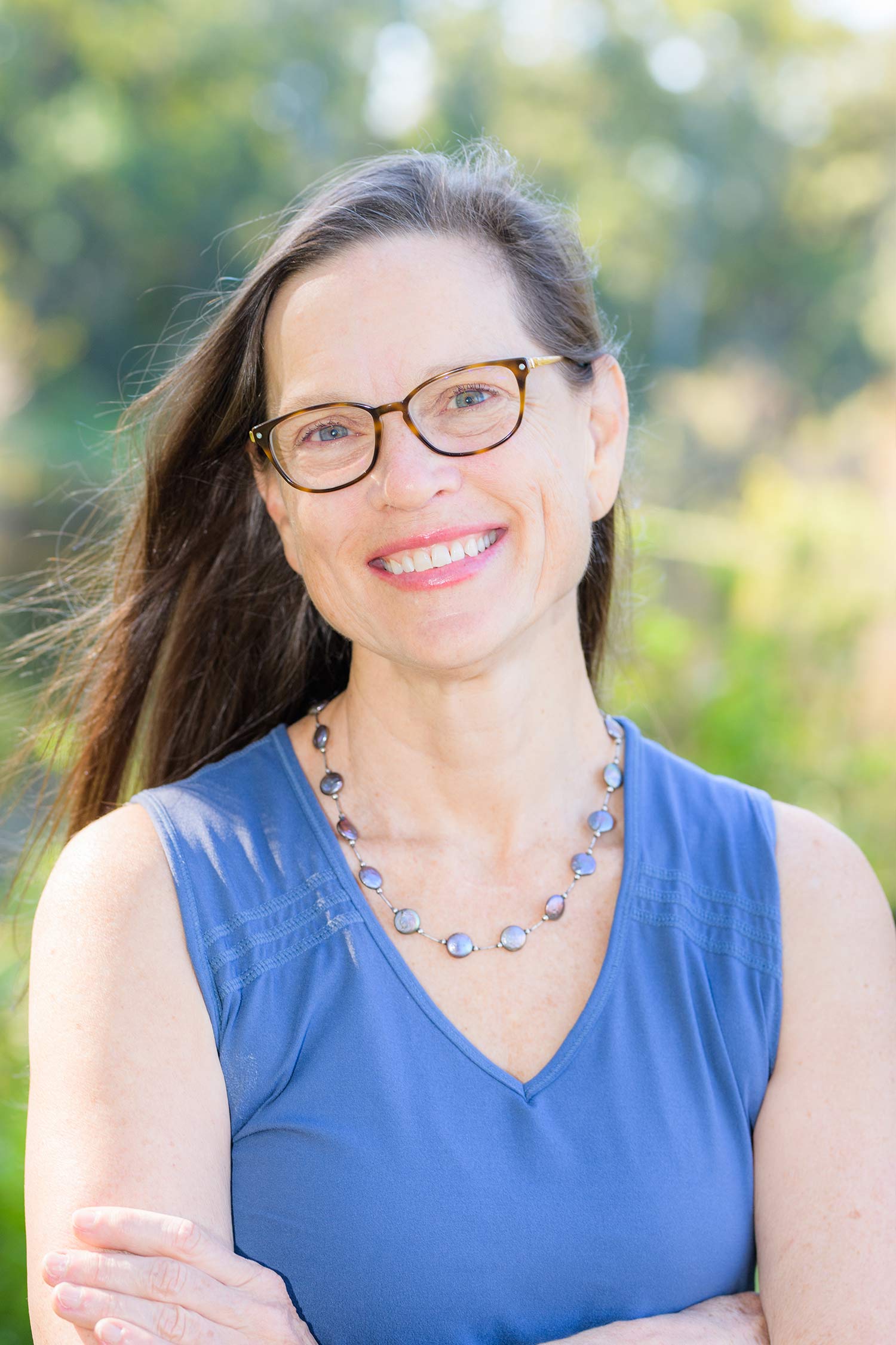 Portrait of a woman smiling outdoors, wearing glasses, a blue sleeveless top and a beaded necklace.