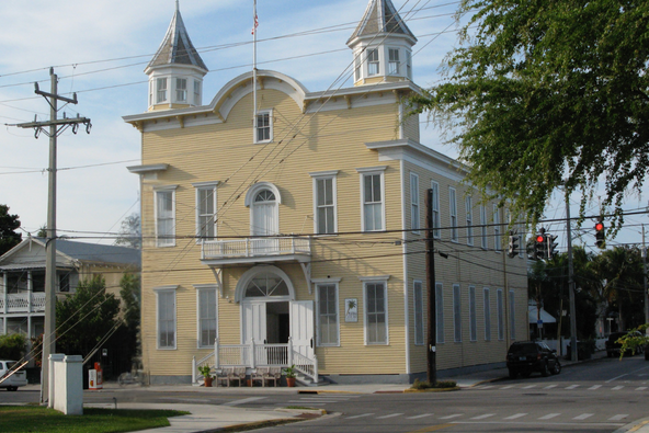 The historic Armory in Key West: a two-story pale yellow clapboard building with twin pointed cupolas, an arched gable, and a second-floor balcony, seen from the street corne
