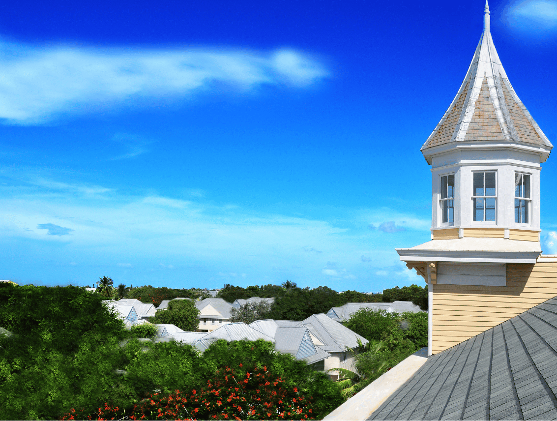 An octagonal white and yellow cupola with a steep shingled spire rises from a gray rooftop, overlooking the tin roofs and treetops of Key West under a deep blue sky.