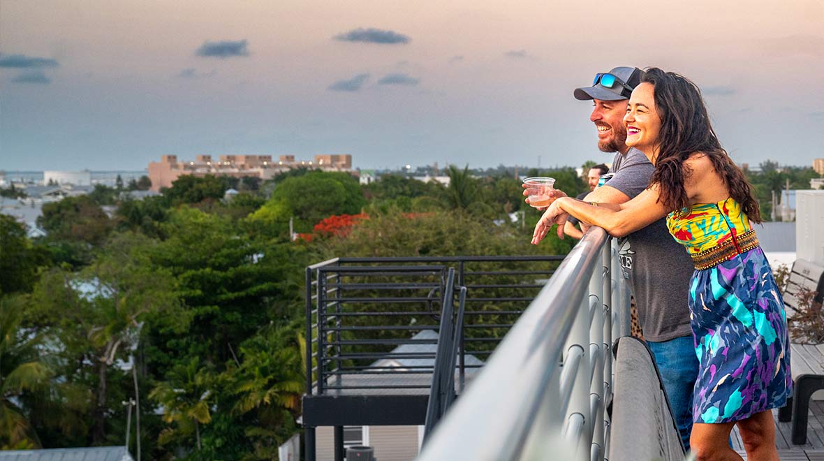 Two guests smile and lean against the railing at Hugh’s View, overlooking the treetops and rooftops of Key West at sunset.