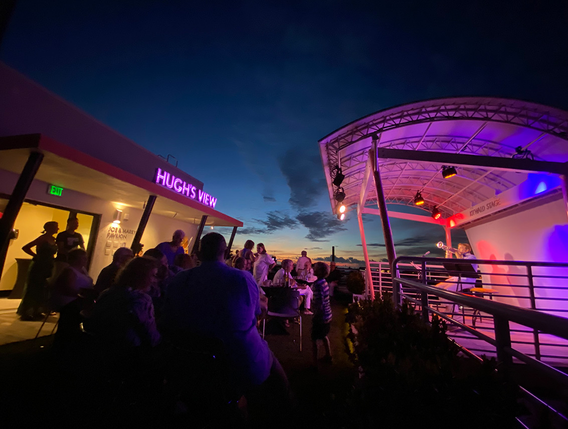 Guests gather on Hugh’s View rooftop during an evening performance, with the stage glowing under colorful lights against the sunset sky.